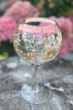 Ett stort gin- och vinglas med blommönster och texten "Bee-autiful friend" står utomhus med rosa blommor i bakgrunden.