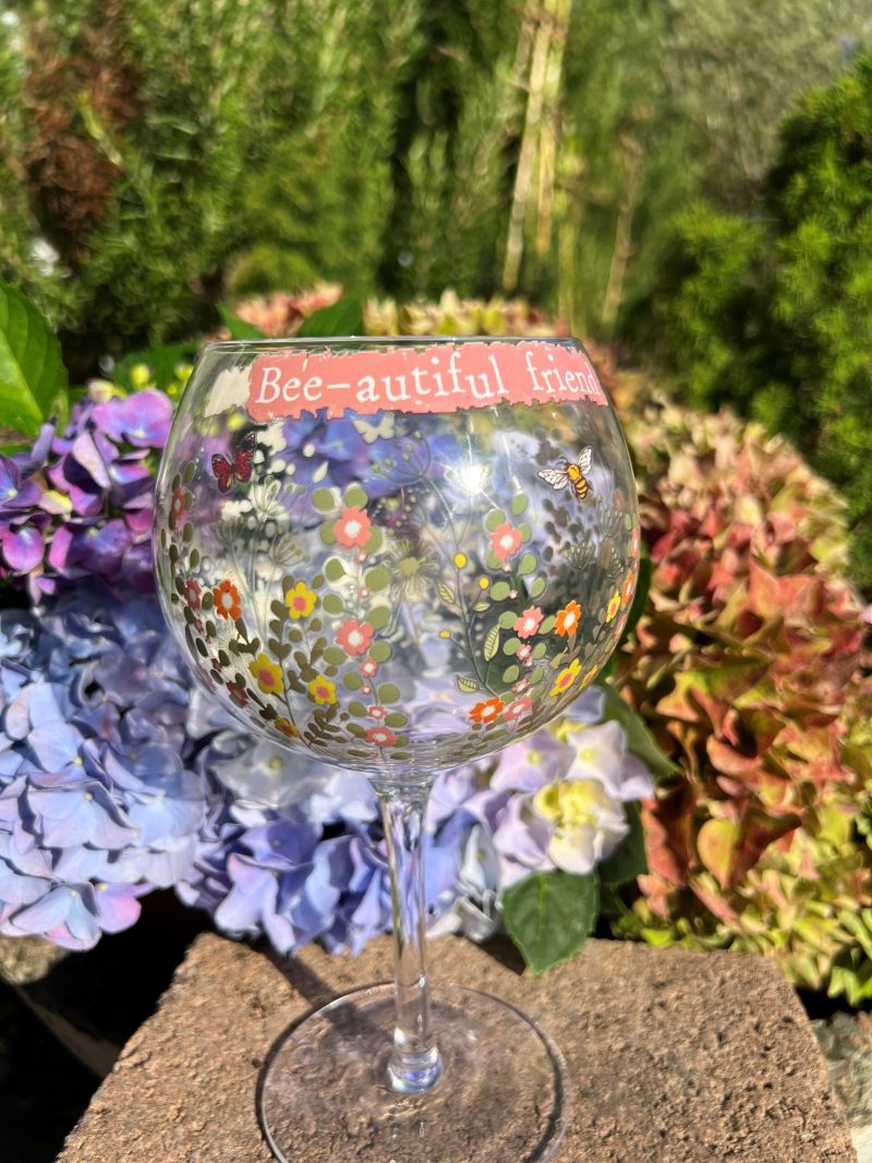 Stort vinglas med blommor, bin och texten "Bee-autiful friend". Glaset står utomhus bland blå och rosa hortensior.