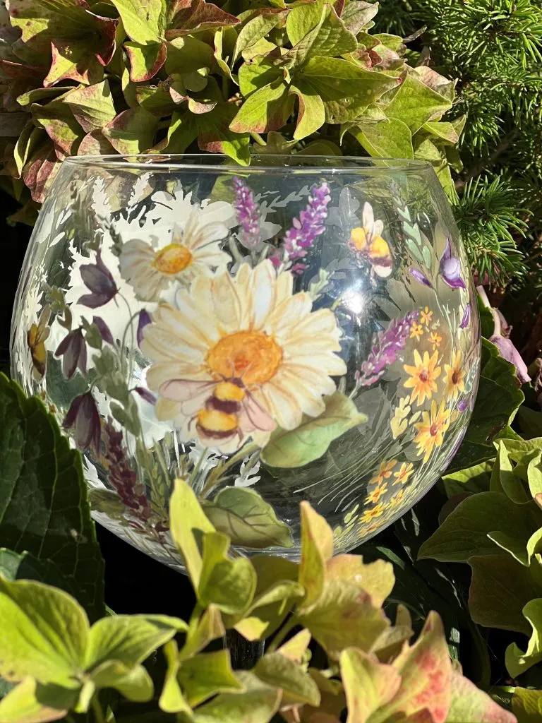 Ett stort gin-glas dekorerat med sommarblommor och bin, placerat utomhus bland gröna blad.