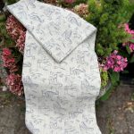 Naturfärgad linneduk med gråblått mönster av ankor och gäss, draperad över en blomkruka utomhus.