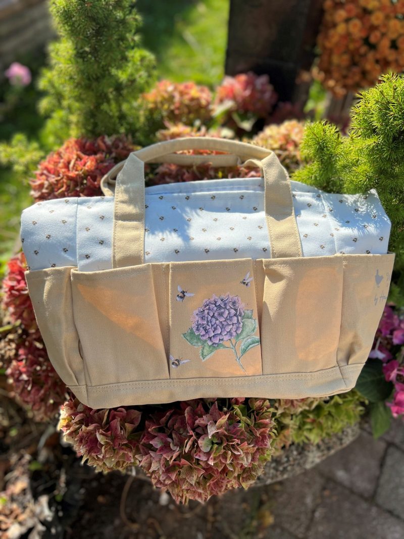 En beige och ljusblå trädgårdsväska med tryck av en lila hortensia och bin, fotograferad utomhus bland blommor.