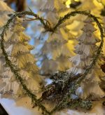 Ett hjärtformat julpynt av en grön kvist ramar in två vita, snötäckta granar med silverglitter.