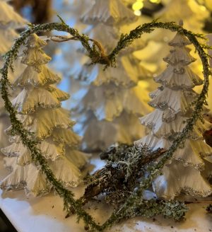 Ett hjärtformat julpynt av en grön kvist ramar in två vita, snötäckta granar med silverglitter.