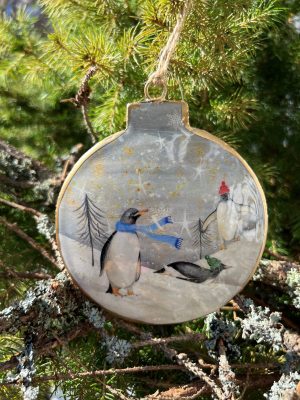 Rund juldekoration med guldkant och motiv av pingviner med mössor och halsduk i ett snöigt landskap, hängandes i en gran.