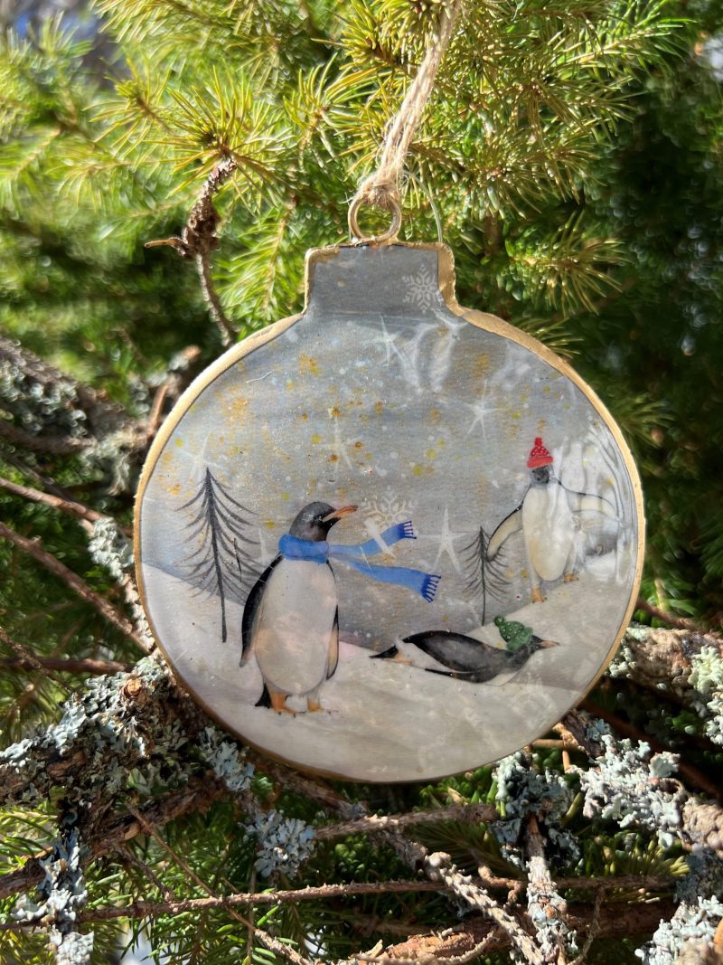 Rund juldekoration med guldkant och motiv av pingviner med mössor och halsduk i ett snöigt landskap, hängandes i en gran.