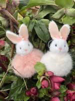 Två fluffiga kaniner, en vit och en rosa, med rosetter i öronen, sitter bland gröna blad och mörkrosa blommor.