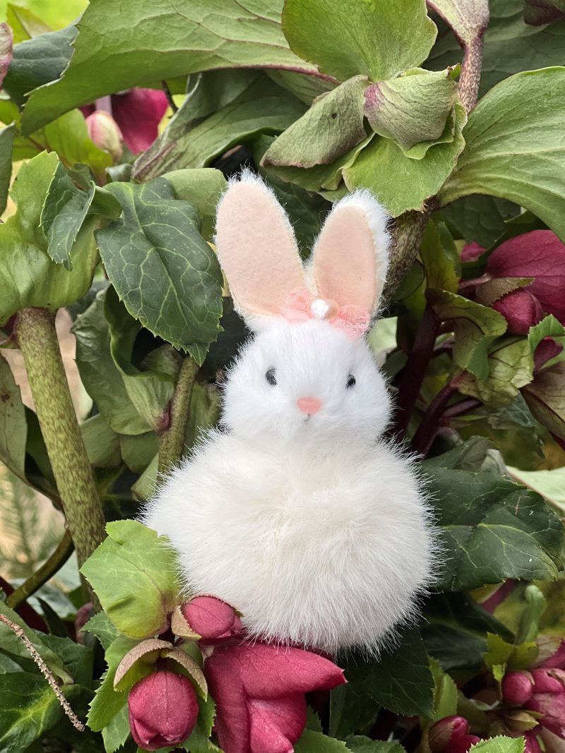 En liten, fluffig vit kaninfigur med rosa öron och rosett, placerad bland gröna blad och mörkrosa blomknoppar.